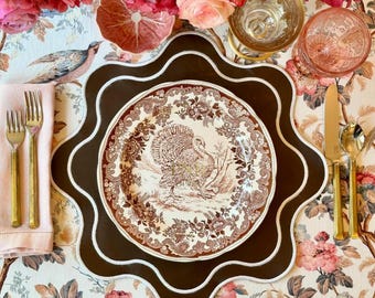 May include: A brown and white ceramic dinner plate with a turkey design. The plate is on a brown scalloped charger with a white border. The charger is on a floral tablecloth.