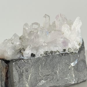 May include: A cluster of clear quartz crystals sits atop a dark gray rock base. The crystals are translucent with hints of purple and white, and they are clustered together in a natural formation. The rock base has a rough, textured surface.