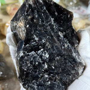 May include: A large, raw, smoky quartz crystal with a dark, almost black color. The surface has a rough, textured appearance with reflective, crystalline facets. The crystal is held in a gloved hand, showcasing its natural form and unique characteristics.