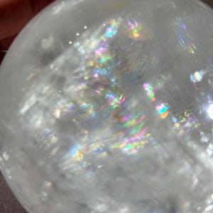 May include: A clear, spherical quartz crystal ball featuring internal fractures and inclusions that create vibrant, multi-colored rainbow light refractions throughout the transparent, polished surface of the stone.