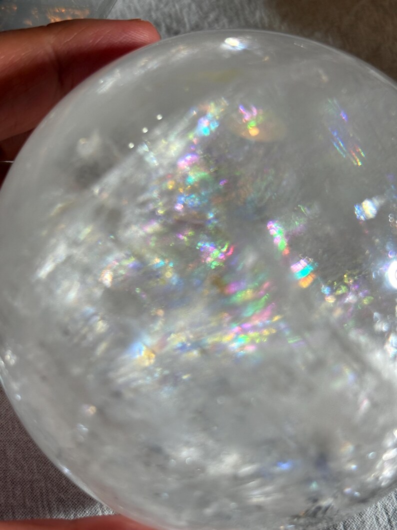 May include: A clear, spherical quartz crystal ball featuring internal fractures and inclusions that create vibrant, multi-colored rainbow light refractions throughout the transparent, polished surface of the stone.