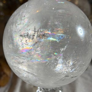 May include: A clear, spherical quartz crystal ball featuring internal fractures and inclusions that create vibrant, multi-colored rainbow light refractions across its polished, smooth surface. The sphere is transparent with visible natural mineral patterns.