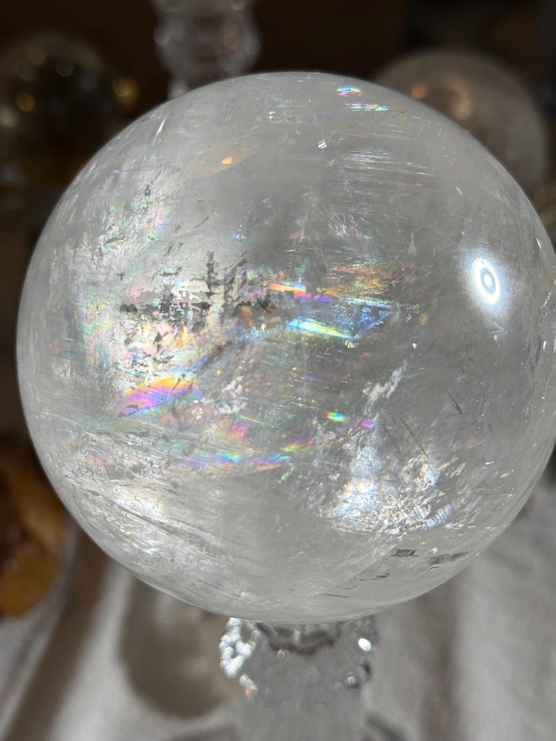 May include: A clear, spherical quartz crystal ball featuring internal fractures and inclusions that create vibrant, multi-colored rainbow light refractions across its polished, smooth surface. The sphere is transparent with visible natural mineral patterns.