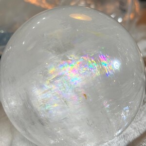 May include: A clear, spherical quartz crystal ball featuring internal fractures that refract light into a vibrant rainbow spectrum. The polished surface is smooth and transparent, showcasing natural inclusions and a distinct prismatic effect.