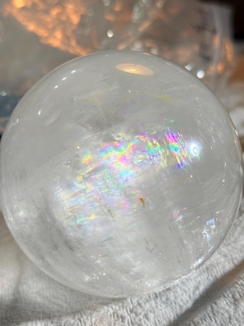 May include: A clear, spherical quartz crystal ball featuring internal fractures that refract light into a vibrant rainbow spectrum. The polished surface is smooth and transparent, showcasing natural inclusions and a distinct prismatic effect.