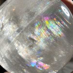 May include: A clear, spherical quartz crystal ball featuring internal fractures and inclusions that create vibrant, multi-colored rainbow light refractions across its polished, smooth surface when illuminated by a bright light source.