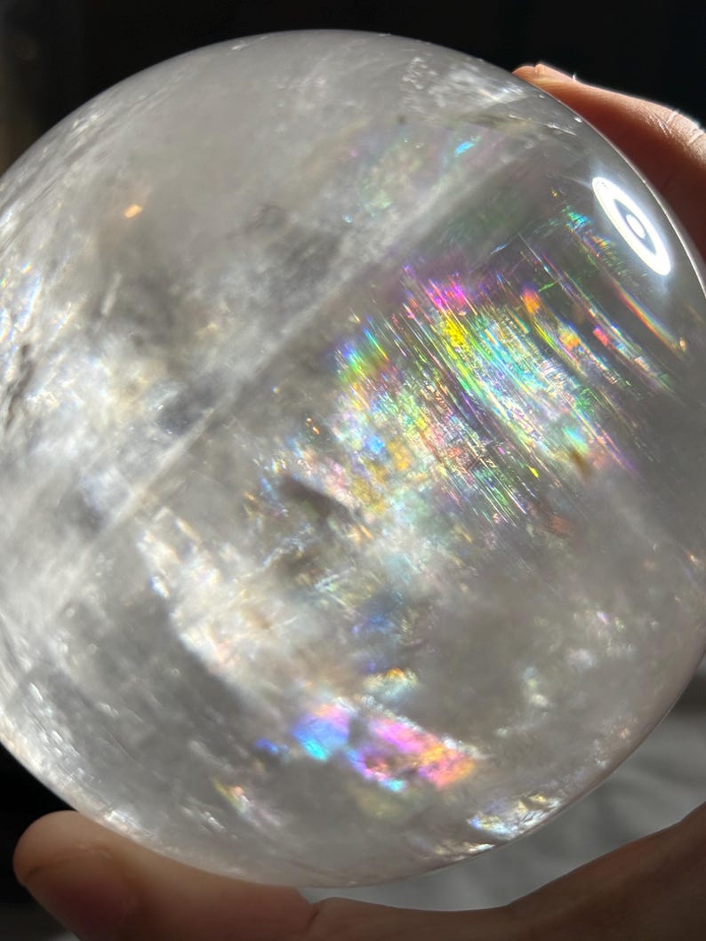 May include: A clear, spherical quartz crystal ball featuring internal fractures and inclusions that create vibrant, multi-colored rainbow light refractions across its polished, smooth surface when illuminated by a bright light source.