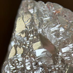 May include: Close-up of a clear, crystalline mineral with a rough, textured surface. The facets reflect light, creating a shimmering effect. The mineral appears to be a natural formation, possibly a quartz or similar crystal.