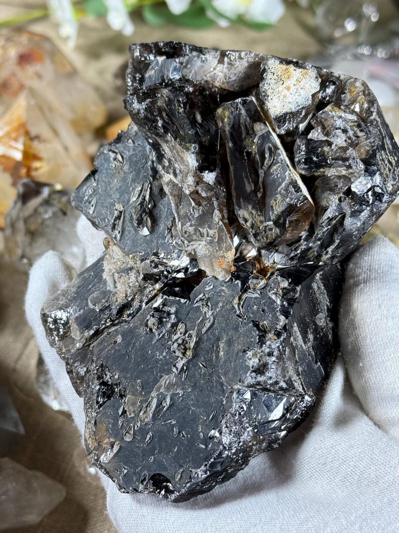 May include: A dark, rough-textured crystal cluster with a mix of black and translucent sections. The crystal has a jagged, irregular shape with reflective surfaces. The overall appearance is of a natural, unpolished mineral specimen.