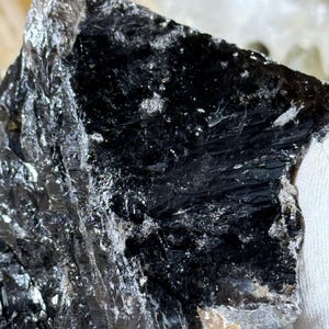 May include: A close-up of a dark, glossy mineral specimen. The stone is primarily black with areas of silver and gray. The surface has a layered, textured appearance, with visible crystalline structures and reflective facets. The overall shape is irregular.