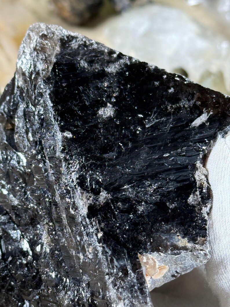 May include: A close-up of a dark, glossy mineral specimen. The stone is primarily black with areas of silver and gray. The surface has a layered, textured appearance, with visible crystalline structures and reflective facets. The overall shape is irregular.
