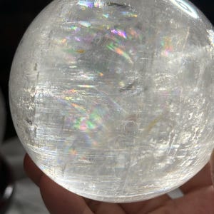 May include: A clear, spherical quartz crystal ball featuring internal fractures and inclusions that create vibrant, multi-colored rainbow light refractions throughout the transparent, polished surface of the stone sphere.