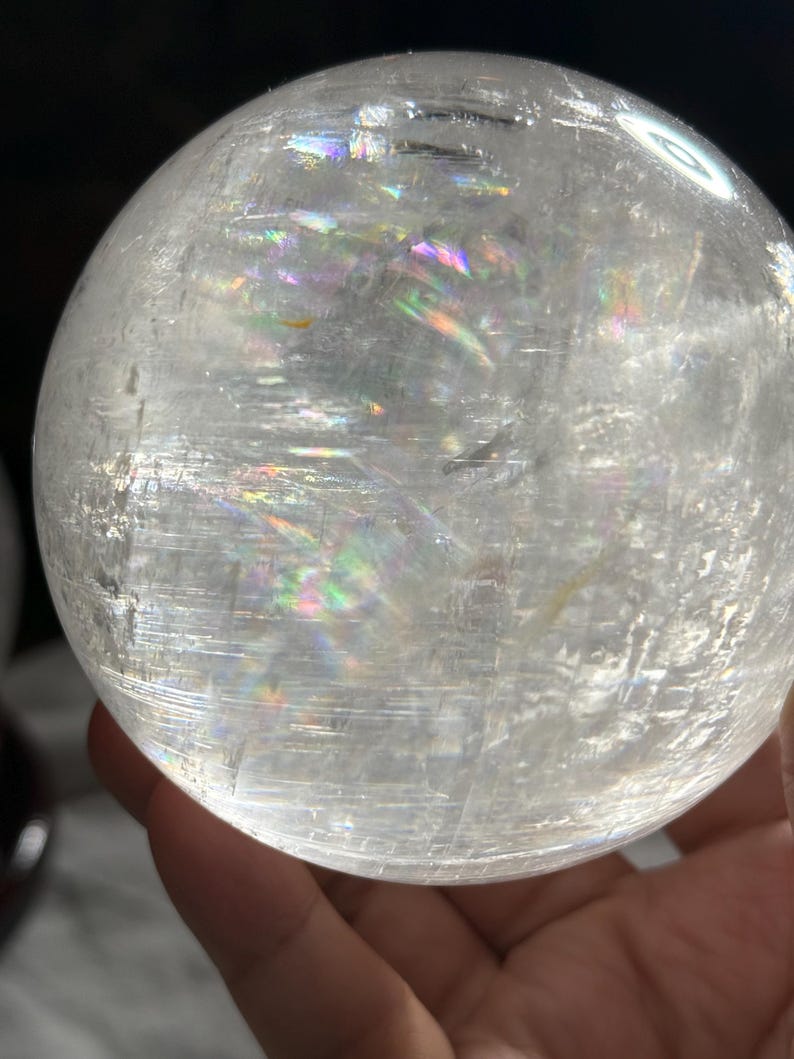 May include: A clear, spherical quartz crystal ball featuring internal fractures and inclusions that create vibrant, multi-colored rainbow light refractions throughout the transparent, polished surface of the stone sphere.