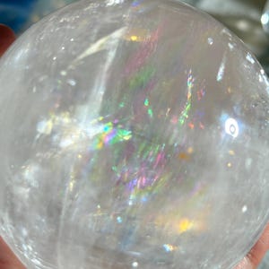 May include: A clear, spherical quartz crystal ball featuring internal fractures that create vibrant, multi-colored rainbow light reflections throughout the transparent stone. The surface is smooth and polished, showcasing natural inclusions.