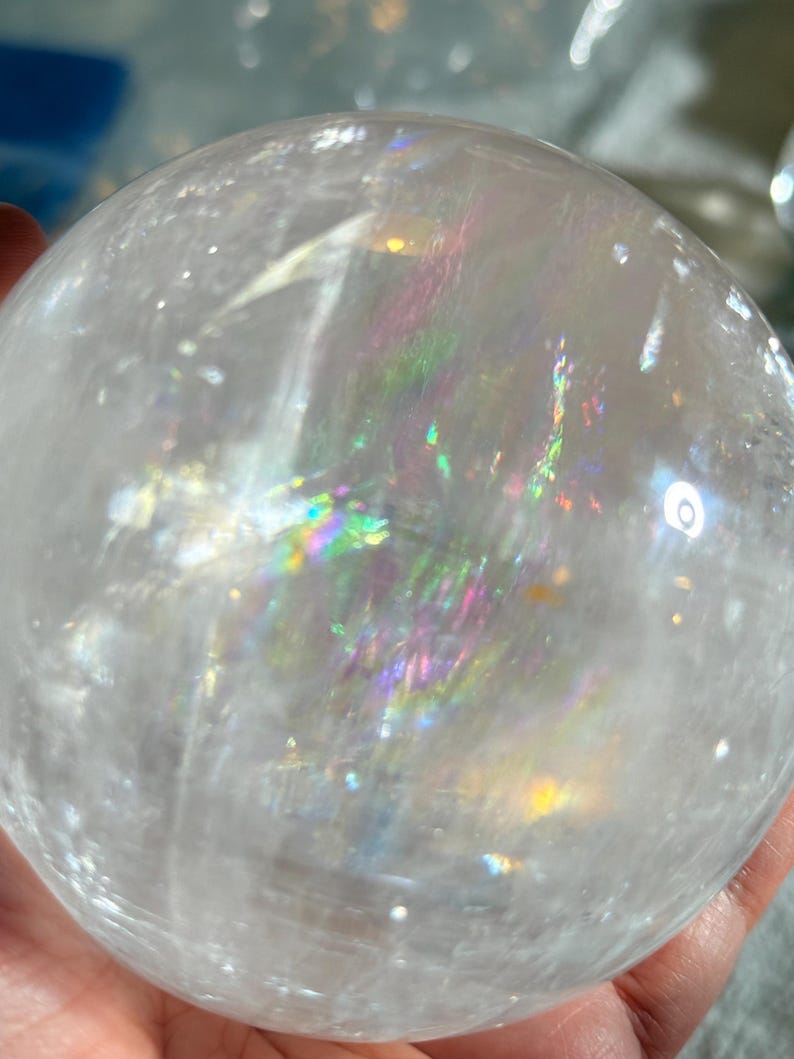 May include: A clear, spherical quartz crystal ball featuring internal fractures that create vibrant, multi-colored rainbow light reflections throughout the transparent stone. The surface is smooth and polished, showcasing natural inclusions.