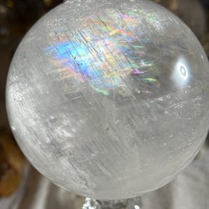 May include: A clear, spherical quartz crystal ball featuring internal fractures and inclusions that create a vibrant, multi-colored rainbow light refraction across the upper surface of the polished, translucent stone sphere.