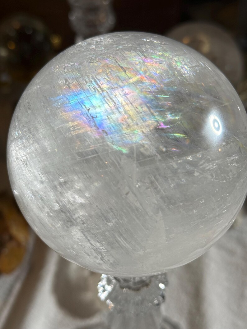 May include: A clear, spherical quartz crystal ball featuring internal fractures and inclusions that create a vibrant, multi-colored rainbow light refraction across the upper surface of the polished, translucent stone sphere.