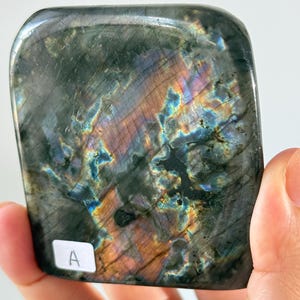 May include: A polished, rectangular labradorite stone with rounded corners, displaying a dark gray base color with iridescent flashes of blue, gold, and purple. A small white square sticker with the black letter A is attached to the surface.