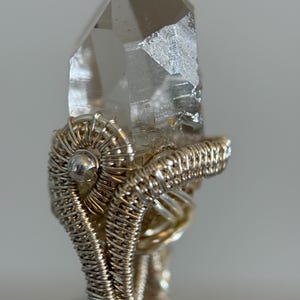 May include: A clear quartz crystal ring with a silver wire wrap. The crystal is a light brown color and has a pointed tip. The wire wrap is intricately designed, creating a unique and artistic piece of jewelry.