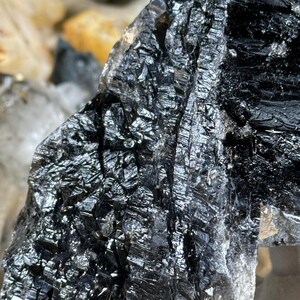 May include: Close-up of a raw, natural black mineral specimen. The surface is highly reflective, with a crystalline structure and a metallic sheen. The mineral has a rough, textured appearance, with visible lines and ridges. The overall color is a deep, lustrous black.