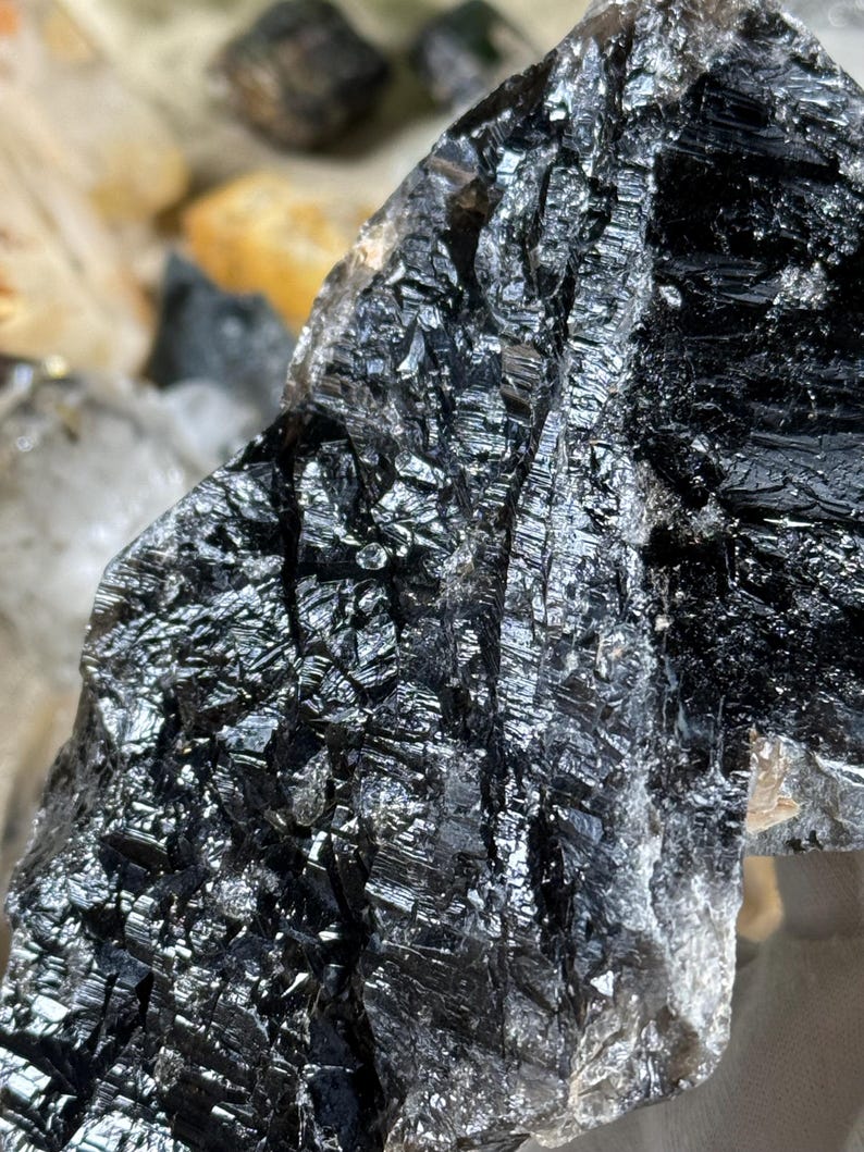 May include: Close-up of a raw, natural black mineral specimen. The surface is highly reflective, with a crystalline structure and a metallic sheen. The mineral has a rough, textured appearance, with visible lines and ridges. The overall color is a deep, lustrous black.