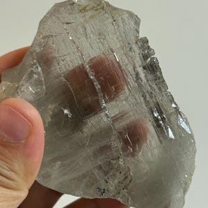 May include: A translucent, irregularly shaped crystal with a rough, textured surface. The crystal is primarily clear with subtle gray and white hues, and some areas appear to reflect light. The overall appearance suggests a natural mineral specimen.