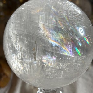 May include: A clear, spherical quartz crystal ball featuring internal fractures and inclusions that create vibrant, multi-colored rainbow light refractions across its polished, smooth surface. The sphere is transparent with visible white streaks.