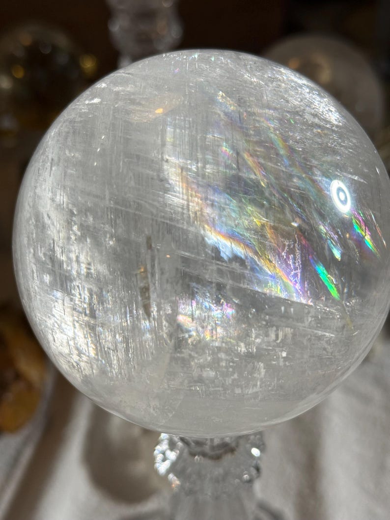 May include: A clear, spherical quartz crystal ball featuring internal fractures and inclusions that create vibrant, multi-colored rainbow light refractions across its polished, smooth surface. The sphere is transparent with visible white streaks.