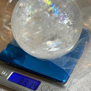 May include: A clear, spherical quartz crystal ball with internal fractures and rainbow light refractions, resting on a digital scale that displays a weight of 881.4 grams. The crystal is partially wrapped in a thin, transparent plastic film.