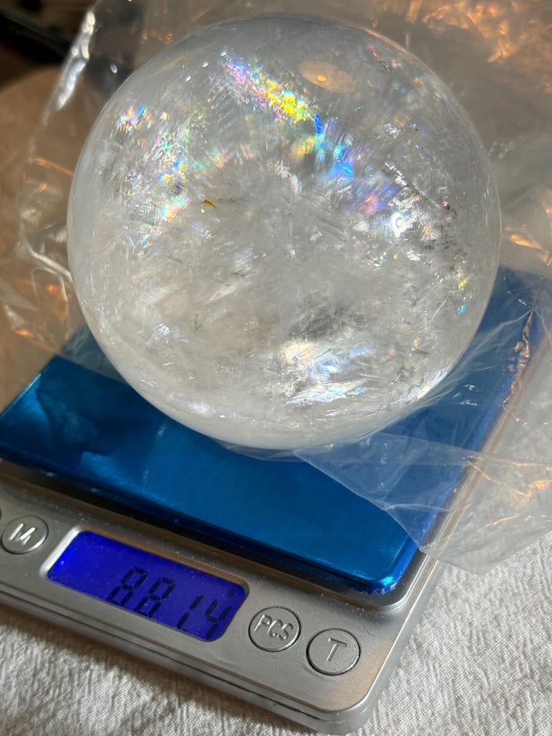 May include: A clear, spherical quartz crystal ball with internal fractures and rainbow light refractions, resting on a digital scale that displays a weight of 881.4 grams. The crystal is partially wrapped in a thin, transparent plastic film.