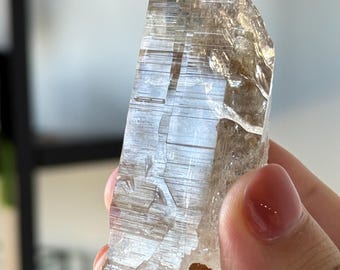 Puede incluir: Una punta de cristal de cuarzo clara y alargada que presenta estrías horizontales distintivas a lo largo de su cuerpo. El espécimen mineral tiene una apariencia translúcida y vítrea con una base rugosa, terrosa y de color marrón.