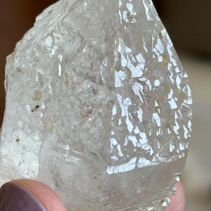 May include: A clear, translucent quartz crystal point featuring a highly textured, faceted surface with numerous small, geometric crystalline structures. The mineral specimen displays natural inclusions and sharp, angular edges throughout its form.
