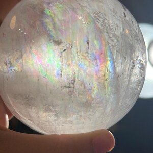 May include: A clear, spherical quartz crystal ball featuring internal fractures and inclusions that create a vibrant, multi-colored rainbow refraction effect across its surface when illuminated by a bright light source.