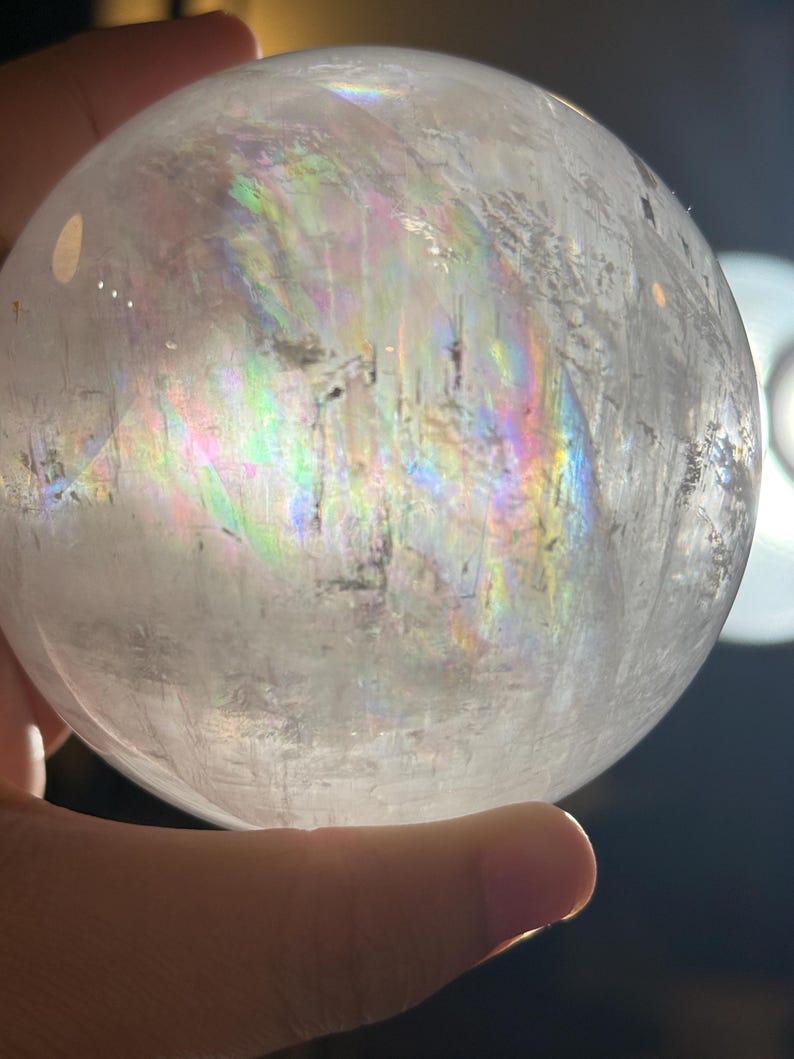 May include: A clear, spherical quartz crystal ball featuring internal fractures and inclusions that create a vibrant, multi-colored rainbow refraction effect across its surface when illuminated by a bright light source.