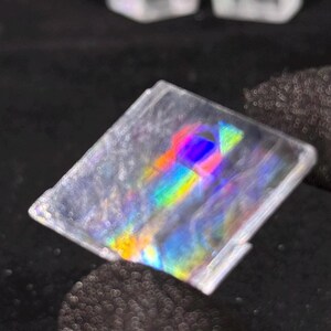 May include: A square, transparent, flat object featuring a central prismatic effect that displays a vibrant spectrum of colours including red, yellow, green, and blue. The surface is clear with a smooth texture, reflecting light to create a rainbow.
