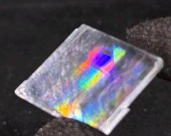 May include: A square, transparent, flat object featuring a central prismatic effect that displays a vibrant spectrum of colors including red, yellow, green, and blue. The surface is clear with a smooth texture, reflecting light to create a rainbow.
