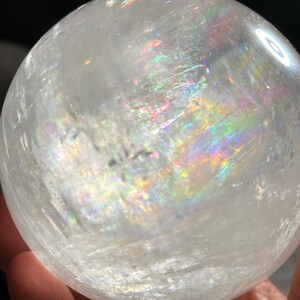 May include: A clear, spherical crystal ball featuring internal fractures and inclusions that create vibrant, multi-colored rainbow light refractions throughout the polished, translucent surface of the stone, showcasing natural mineral patterns.