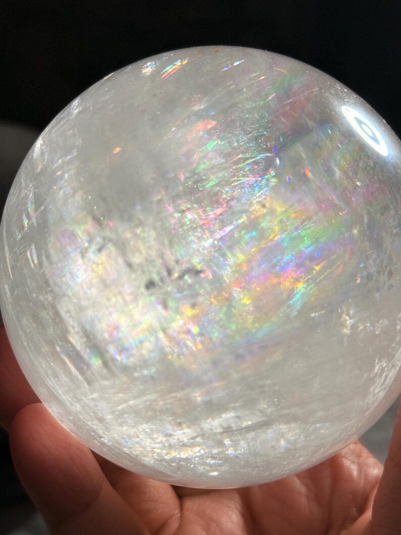 May include: A clear, spherical crystal ball featuring internal fractures and inclusions that create vibrant, multi-colored rainbow light refractions throughout the polished, translucent surface of the stone, showcasing natural mineral patterns.