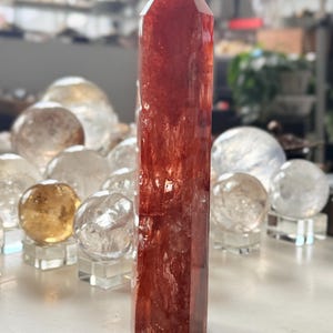 May include: A tall, reddish-orange crystal obelisk with a pointed top. The obelisk is the focal point, with several clear and golden crystal spheres in the background. The crystal has a polished, translucent appearance.