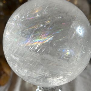 May include: A clear, spherical quartz crystal ball featuring internal fractures and inclusions that create visible rainbow light refractions across its surface. The polished stone has a smooth, rounded texture with a translucent appearance.
