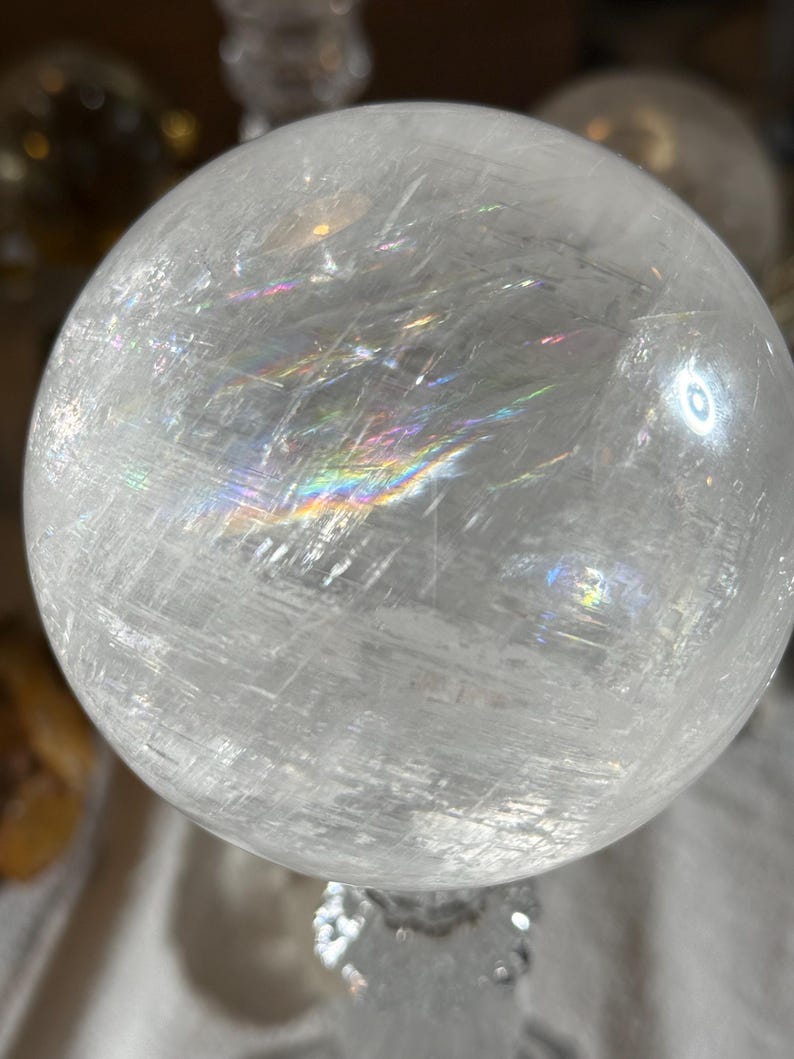 May include: A clear, spherical quartz crystal ball featuring internal fractures and inclusions that create visible rainbow light refractions across its surface. The polished stone has a smooth, rounded texture with a translucent appearance.