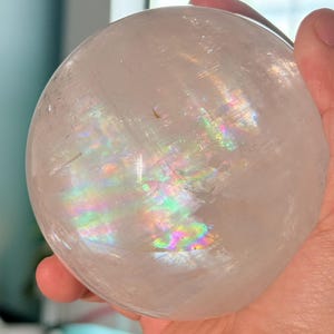 May include: A clear, spherical quartz crystal ball featuring internal fractures that create a vibrant, multi-colored rainbow refraction effect within the stone. The surface of the polished sphere is smooth and transparent, showing natural inclusions.