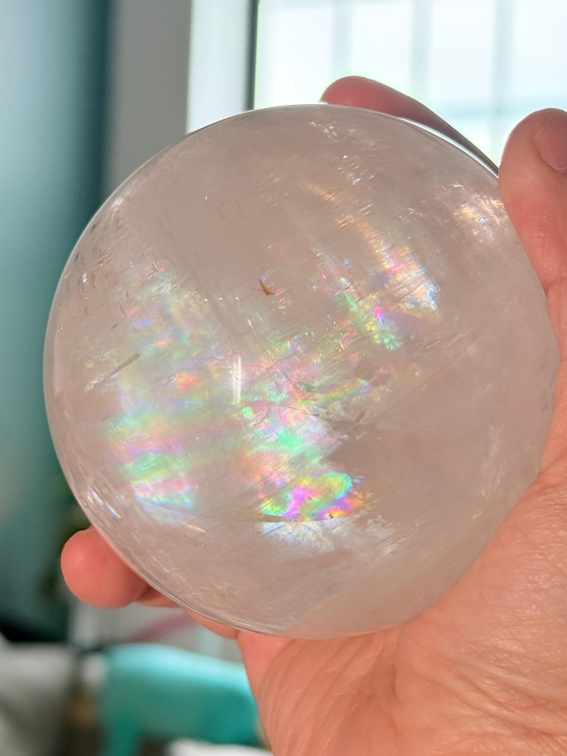 May include: A clear, spherical quartz crystal ball featuring internal fractures that create a vibrant, multi-colored rainbow refraction effect within the stone. The surface of the polished sphere is smooth and transparent, showing natural inclusions.