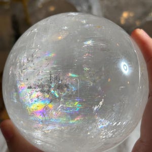May include: A clear, spherical quartz crystal ball featuring internal fractures and inclusions that create vibrant, multi-colored rainbow light refractions across its polished, smooth surface, showcasing natural mineral patterns within the stone.
