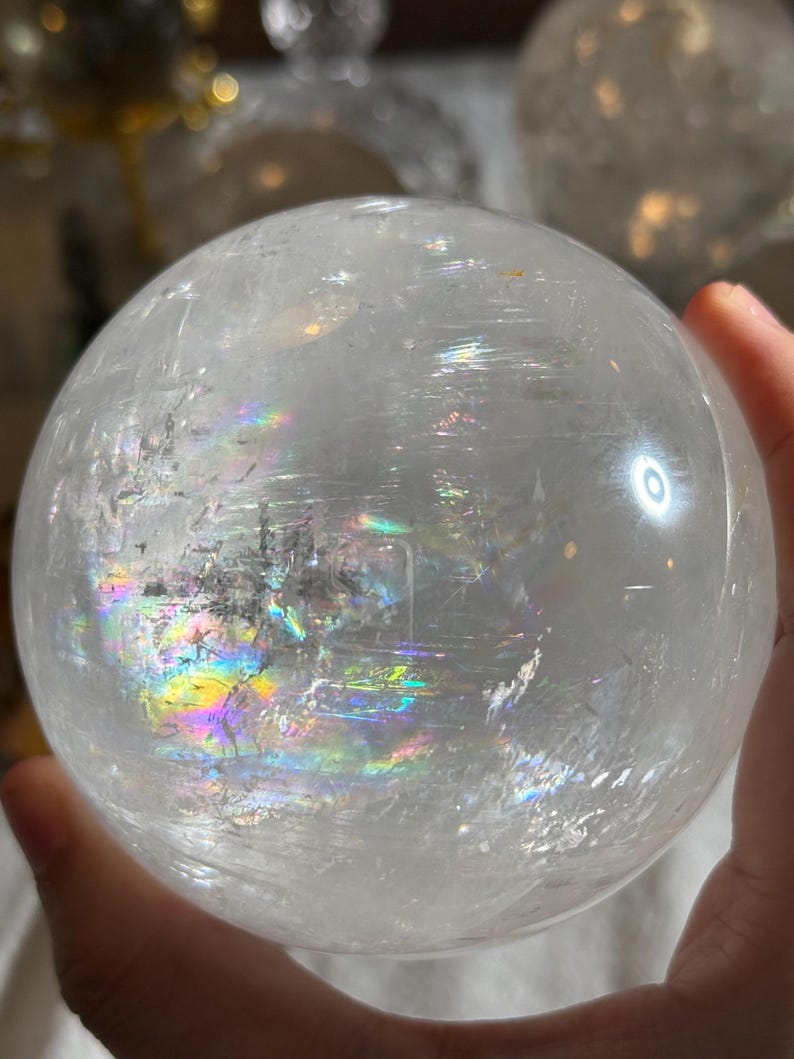 May include: A clear, spherical quartz crystal ball featuring internal fractures and inclusions that create vibrant, multi-colored rainbow light refractions across its polished, smooth surface, showcasing natural mineral patterns within the stone.