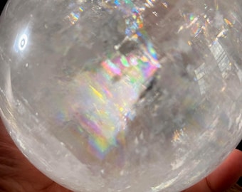 May include: A large, spherical clear quartz crystal ball featuring internal fractures and inclusions that create vibrant, multi-colored rainbow light refractions throughout the transparent, polished stone surface.