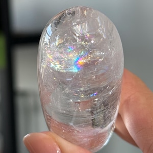 May include: A smooth, oval-shaped clear quartz crystal with a polished surface. The interior of the stone features visible natural fractures and inclusions that create a vibrant, multi-colored rainbow light refraction effect within the transparent body.