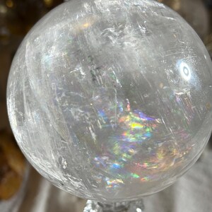 May include: A clear, spherical quartz crystal ball featuring internal fractures and inclusions that create vibrant, multi-colored rainbow light refractions throughout the stone. The surface is smooth and polished, reflecting light from the environment.