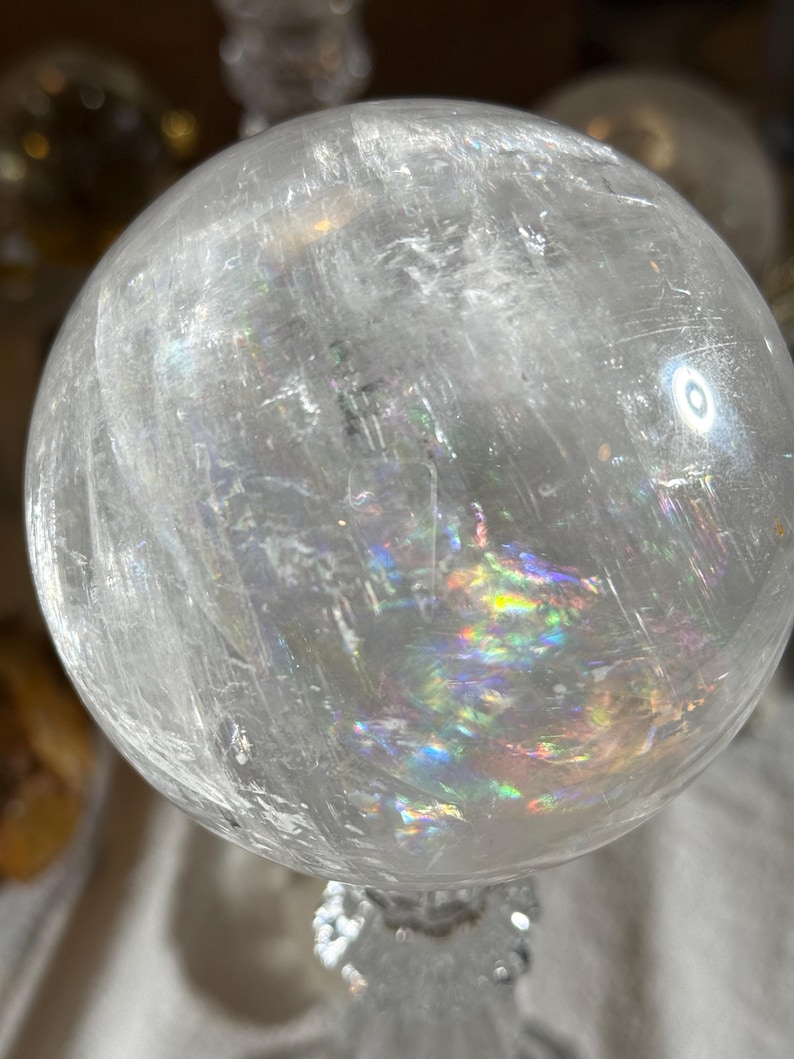 May include: A clear, spherical quartz crystal ball featuring internal fractures and inclusions that create vibrant, multi-colored rainbow light refractions throughout the stone. The surface is smooth and polished, reflecting light from the environment.