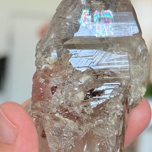 May include: A translucent, smoky quartz crystal specimen featuring a prominent, smooth, flat termination point with a visible internal rainbow refraction. The lower section of the crystal displays a rough, jagged, and textured surface with inclusions.